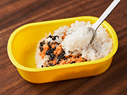 1膳分の白ご飯の半分だけをお弁当箱に詰め、サケフレーク(1人分の半量)・炒りゴマ・揉みのりにしたのりを広げ、残りのご飯をのせてサンドする。